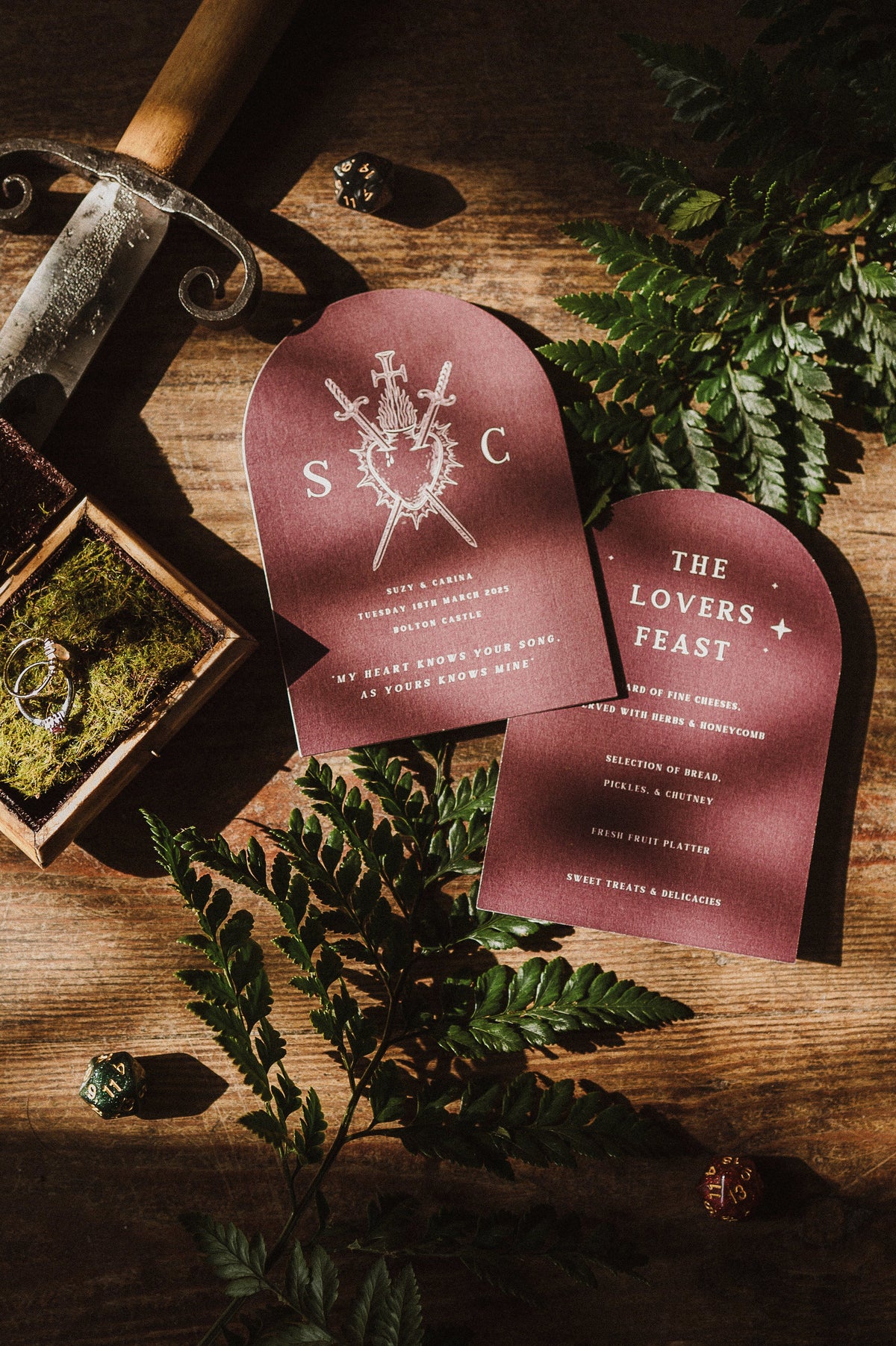 Luxury gothic wedding stationery with arched menus, dramatic burgundy invites, moss ring box, dagger and ferns on rustic wood.