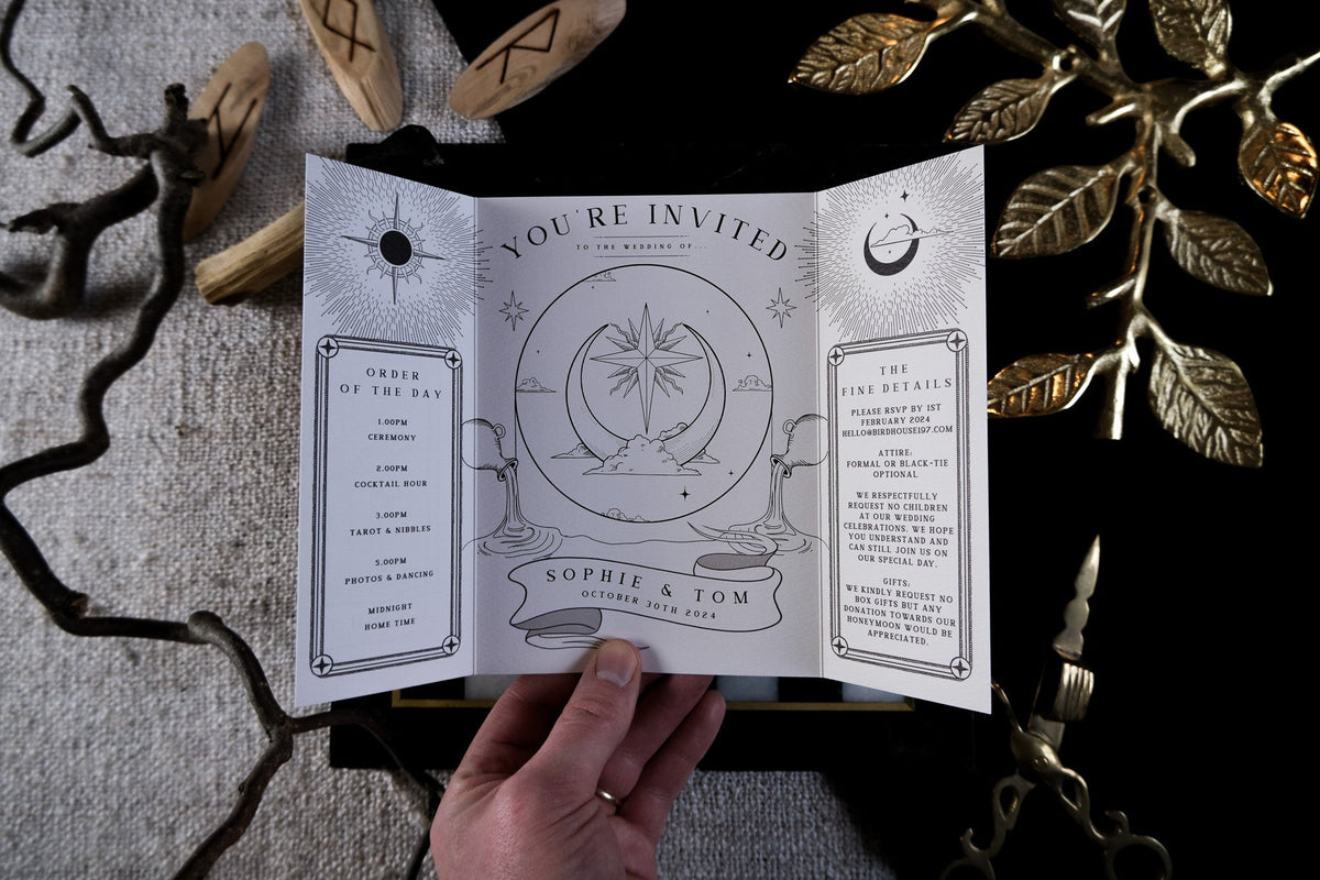 A hand holds a luxury celestial gothic gatefold wedding invitation, featuring tarot-inspired design amid gold leaves and wooden runes.