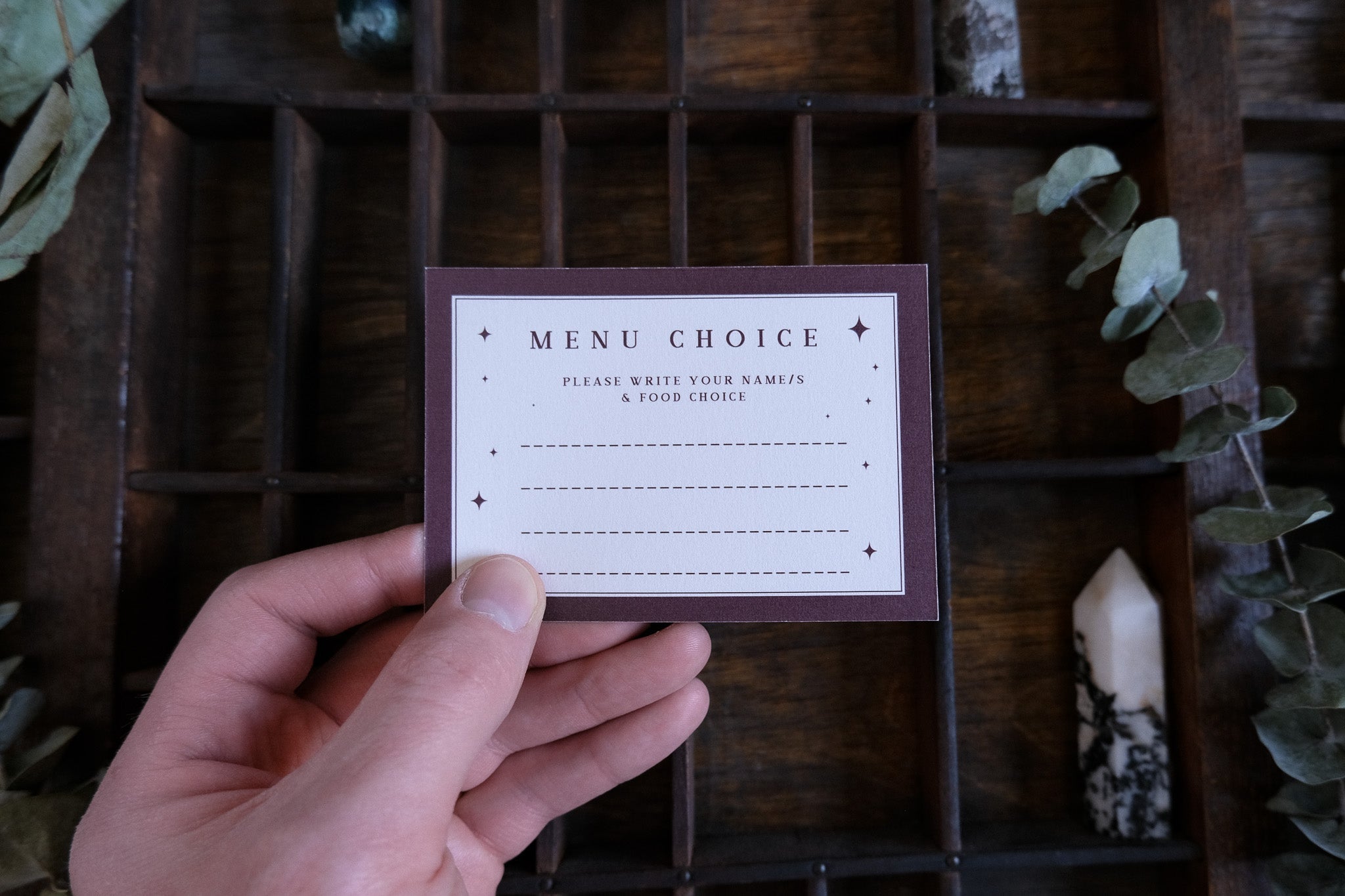 A hand displays a luxury gothic wedding RSVP card with mystical details, set on wood among leaves and UK alternative stationery.