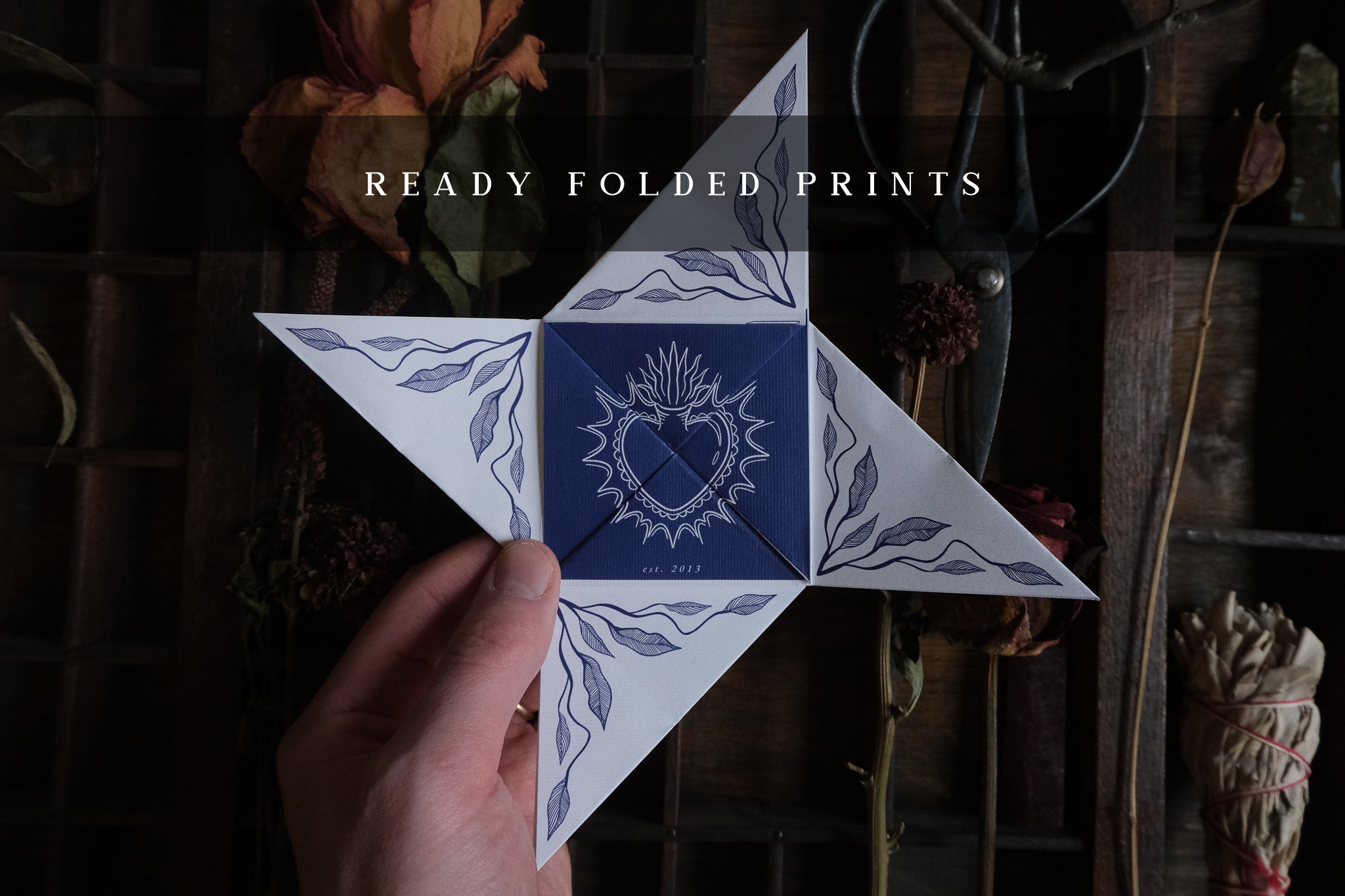 A hand displays a celestial save-the-date wedding invitation with vintage blue and white leaf motifs. Dried flowers and scissors add romance.