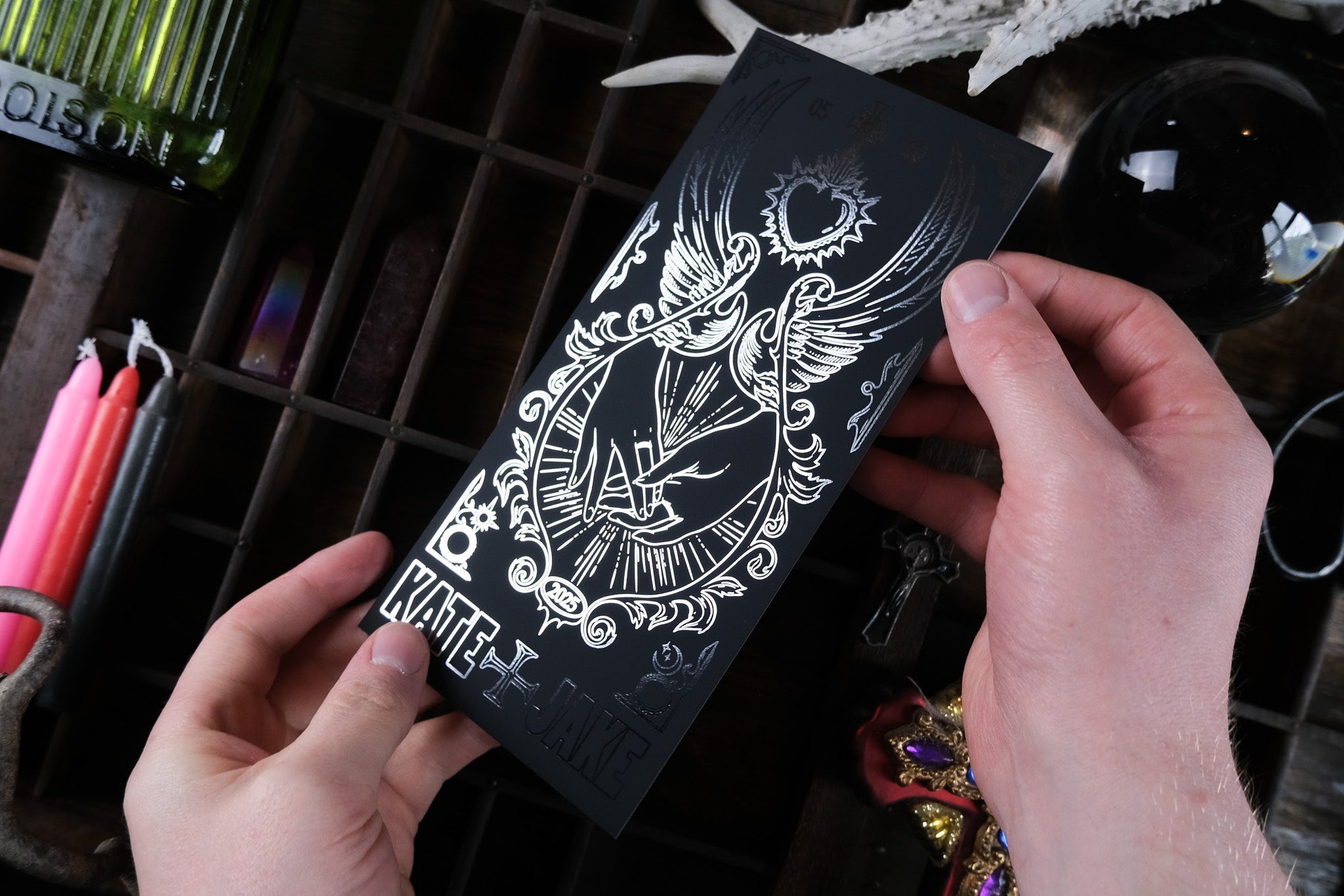 A luxurious, foiled black save the date card with silver gothic hand motifs; dramatic, romantic UK wedding stationery. Candles behind.