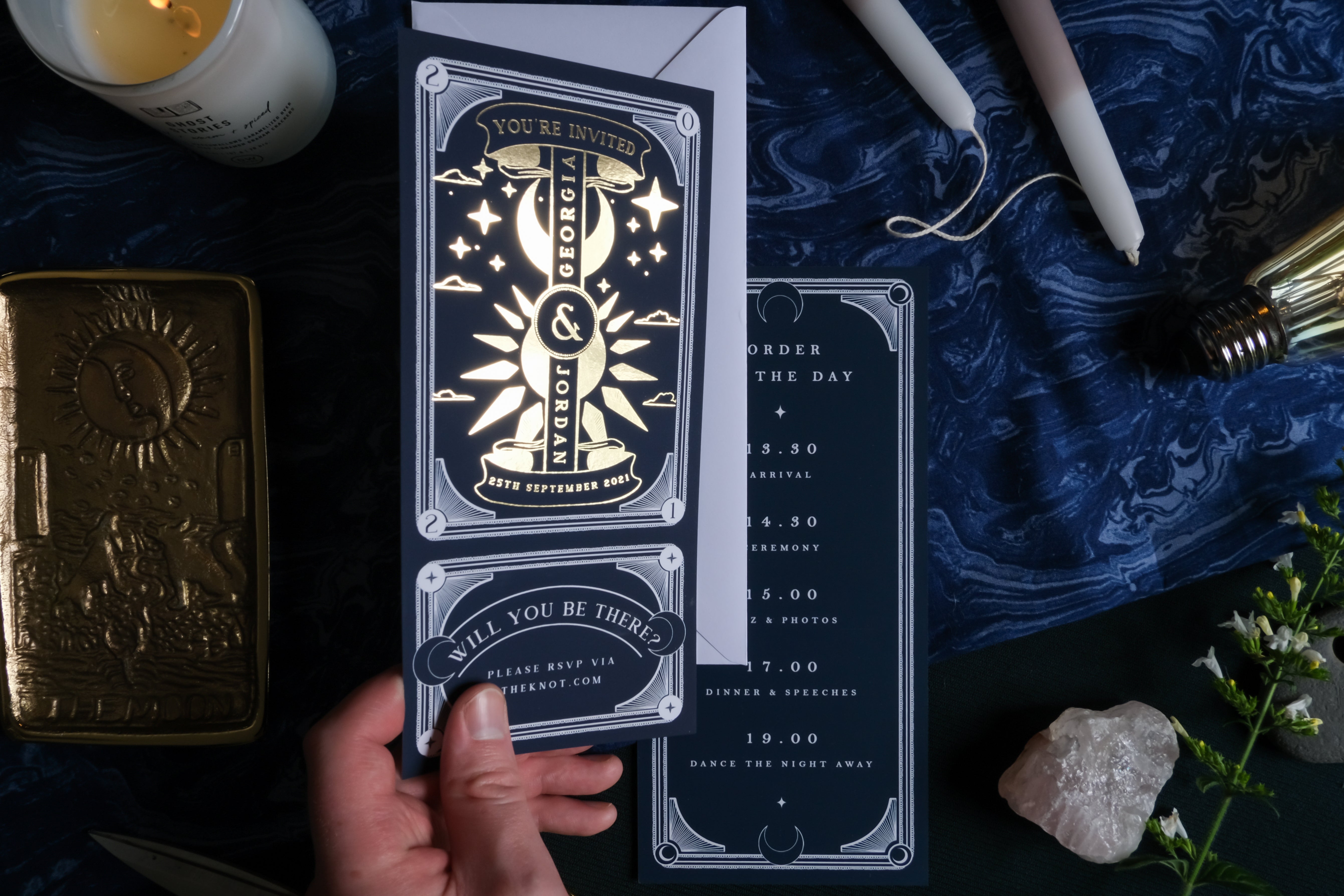 A hand presents luxury celestial wedding stationery with gold foil, set amidst candles, crystals, and florals for a mystical look.