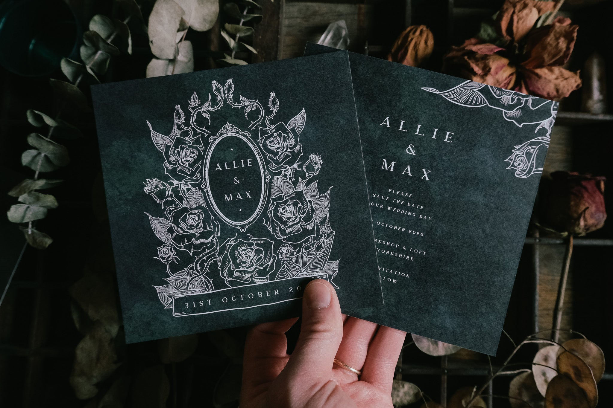 A hand presents two gothic luxury save the date cards with dark floral Victorian design, set against dried flowers; UK wedding stationery.