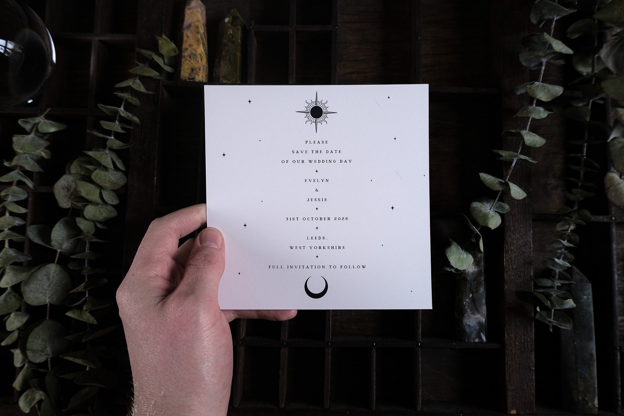 A hand displays a luxury foiled save the date with dramatic celestial motifs, styled amidst dried greenery on rustic wood.