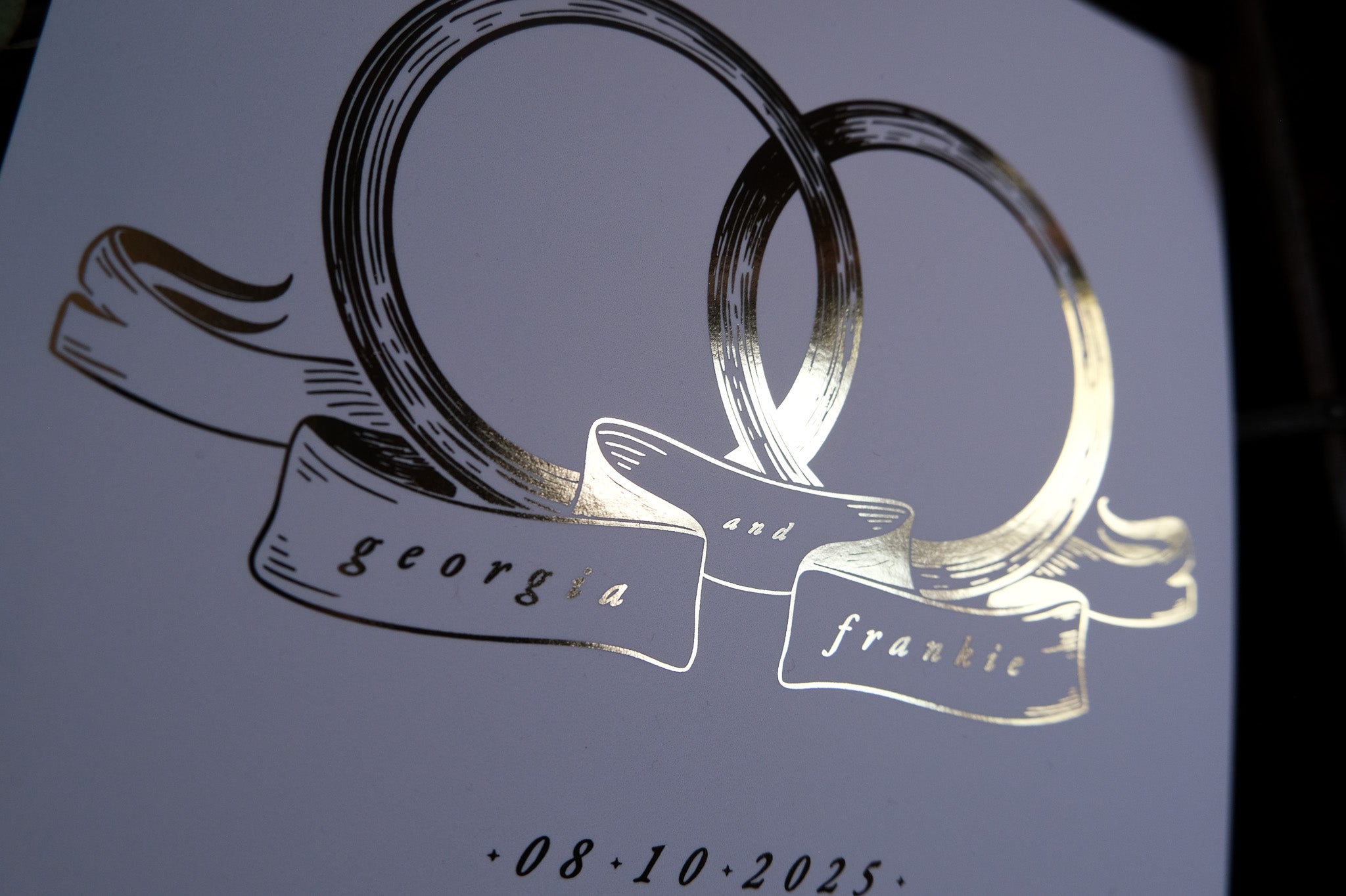 Close-up of luxury foiled Save the Date card, adorned with interlocking gold bands and elegant ribbon banner on premium wedding stationery.