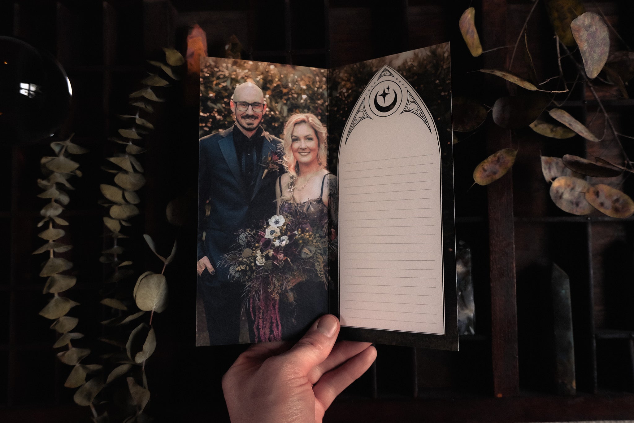 A hand holds a luxury personalised wedding thank you card with gothic lined design, romantic photo, and dried leaves in a dramatic setting.