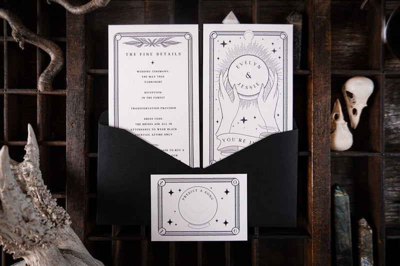 Luxury UK wedding stationery suite with black-and-white celestial, gothic designs, partially revealed from a black envelope on rustic wood.