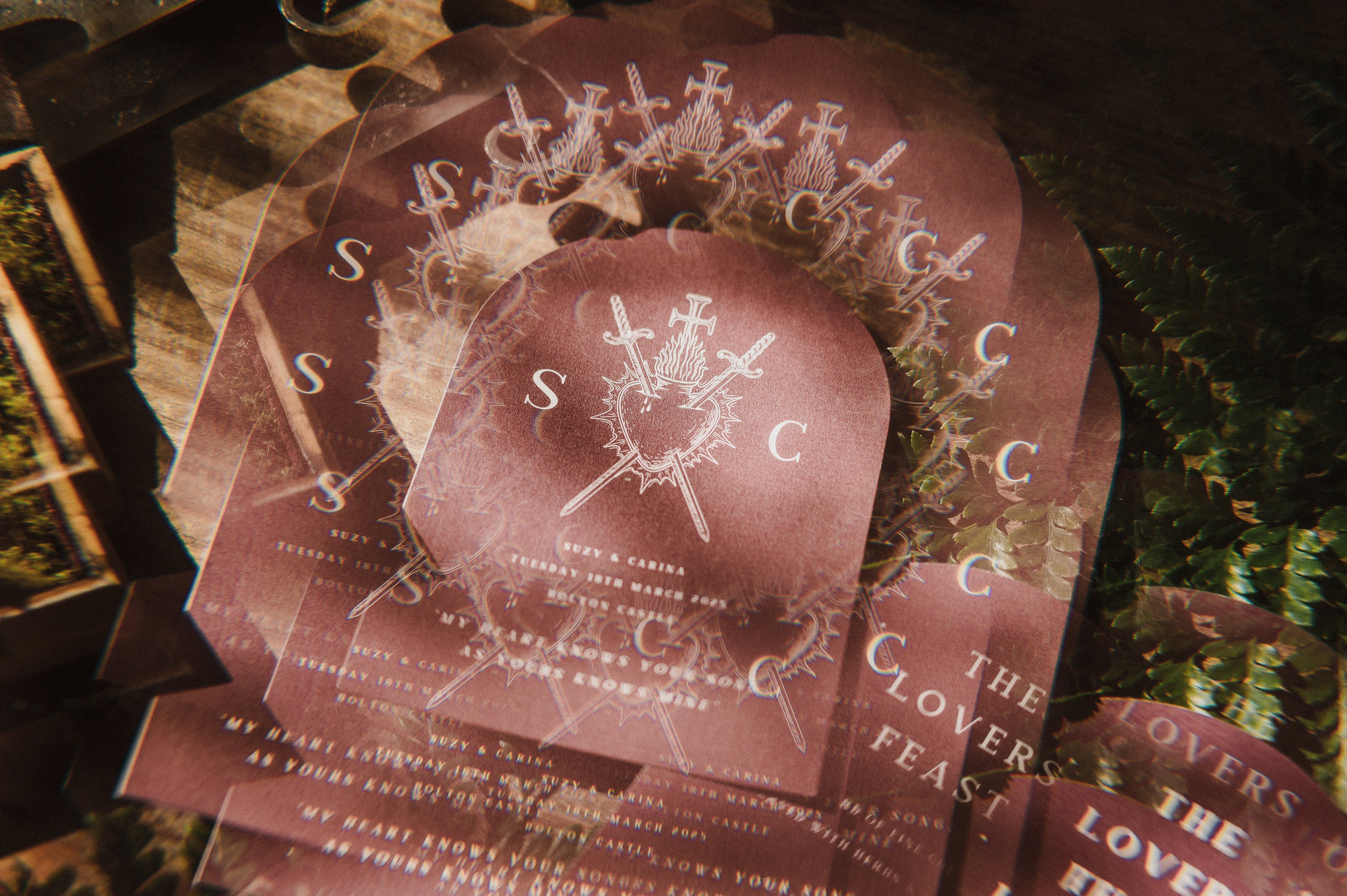 A stack of luxury maroon arched wedding menus with a dramatic white bleeding heart and crossed swords, styled with boho accents.