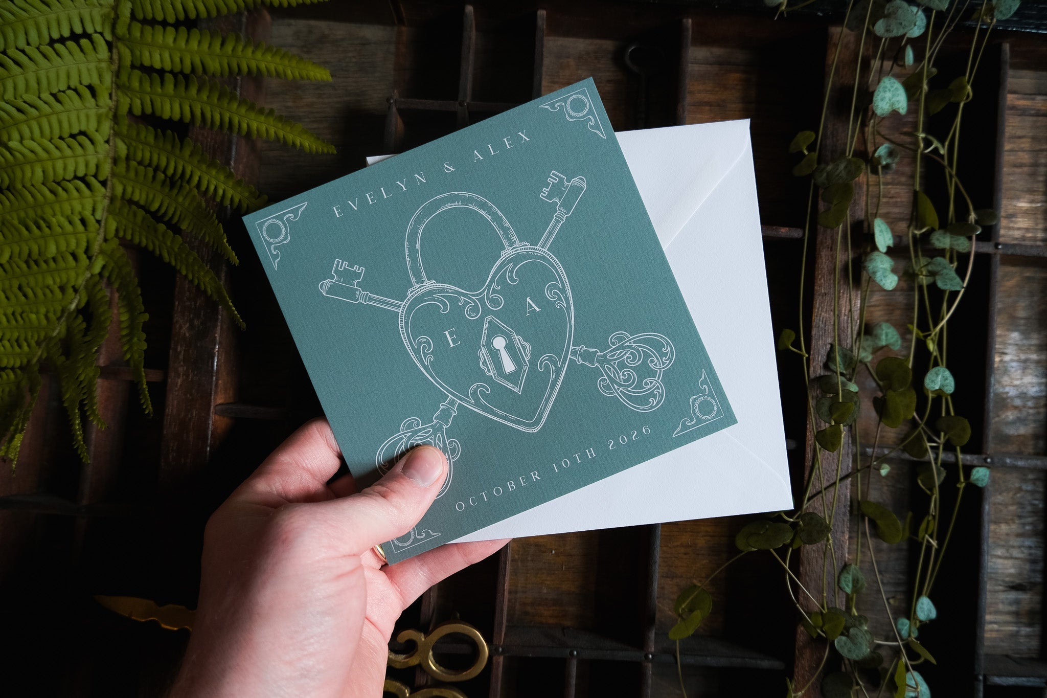 A hand holds a vintage-inspired luxury save the date card with heart padlock and skeleton key motifs, set among foliage and antique scissors.