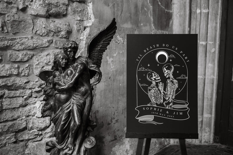A dramatic black-and-white photo features a gothic angel statue beside a mystical tarot-inspired luxury wedding welcome sign with skeleton art.