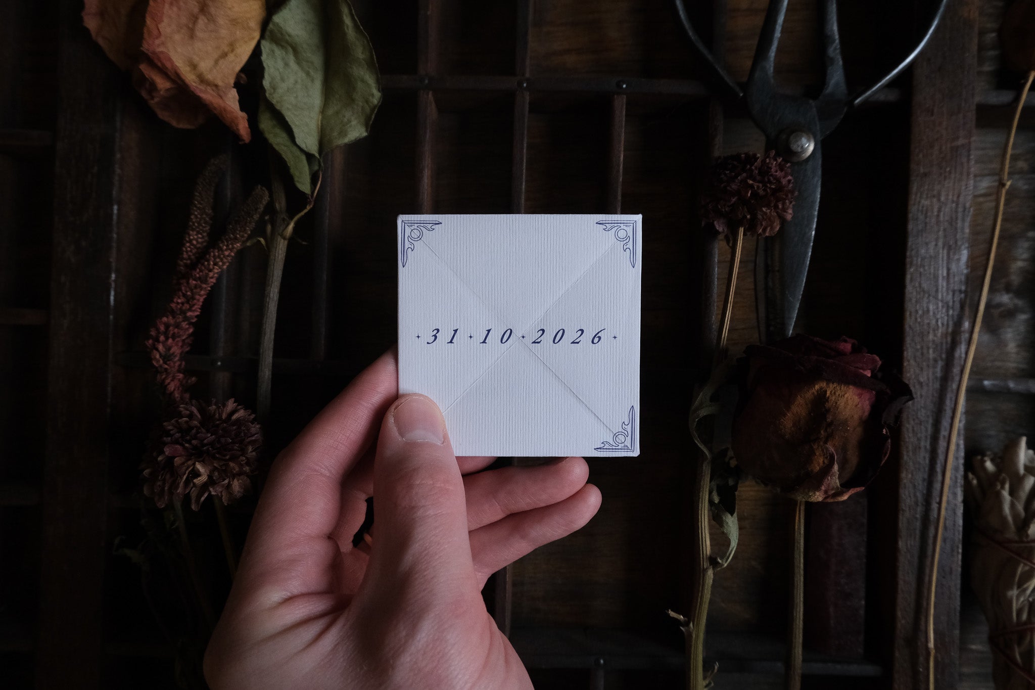 A hand holds a luxury celestial folding save the date with vintage, gothic accents, surrounded by dried flowers and boho wedding stationery.