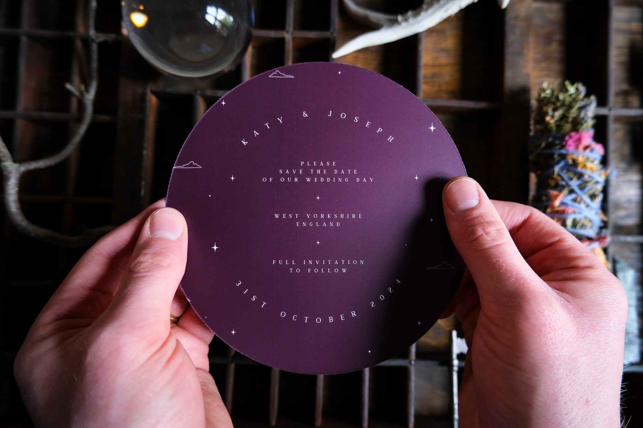 A person holds a luxury round burgundy gothic save the date card with celestial constellation details, perfect for alternative UK weddings.