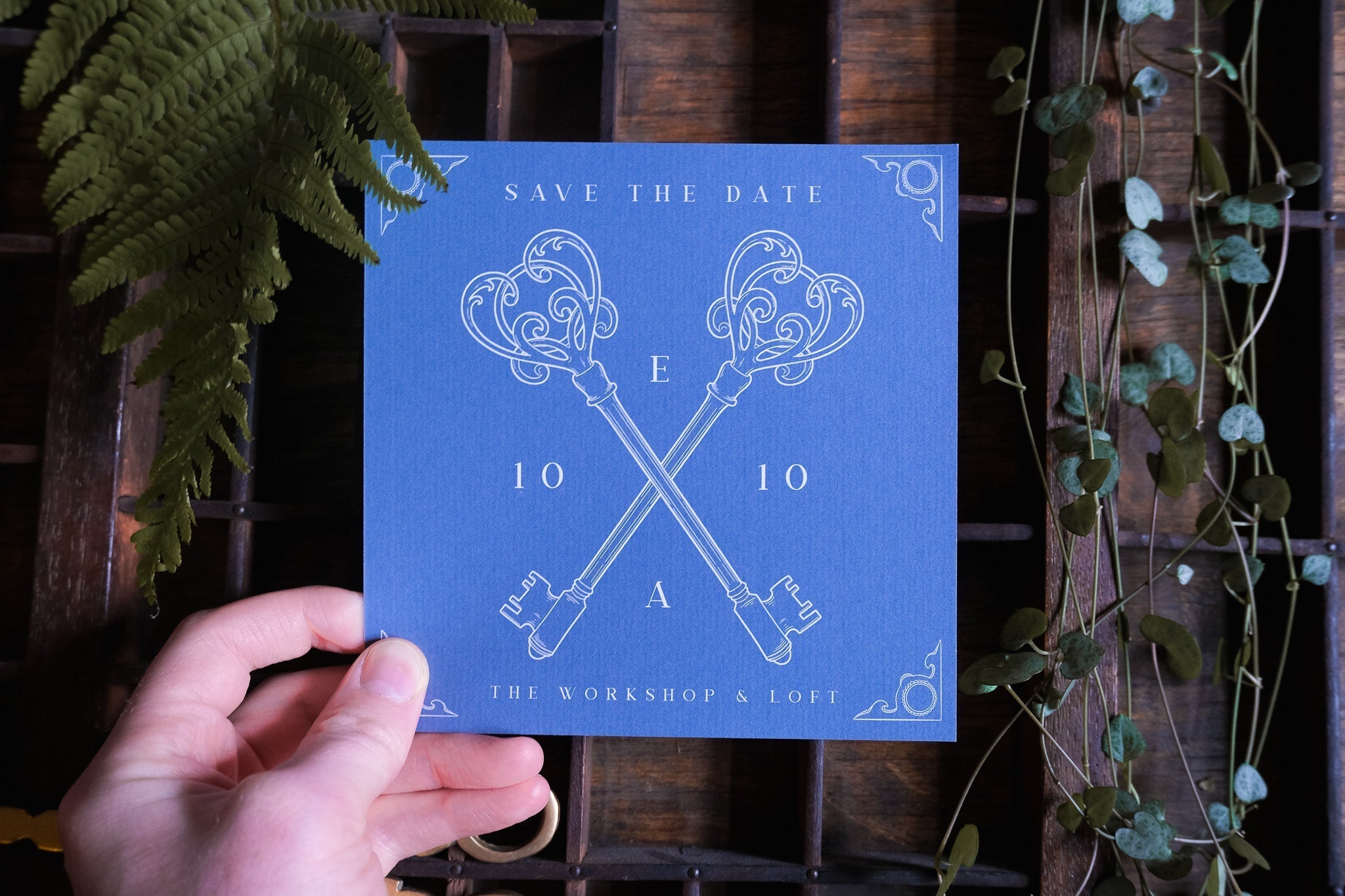 A hand holds a luxury vintage-inspired save the date card with crossed skeleton keys, Victorian details, and botanical accents on wood.