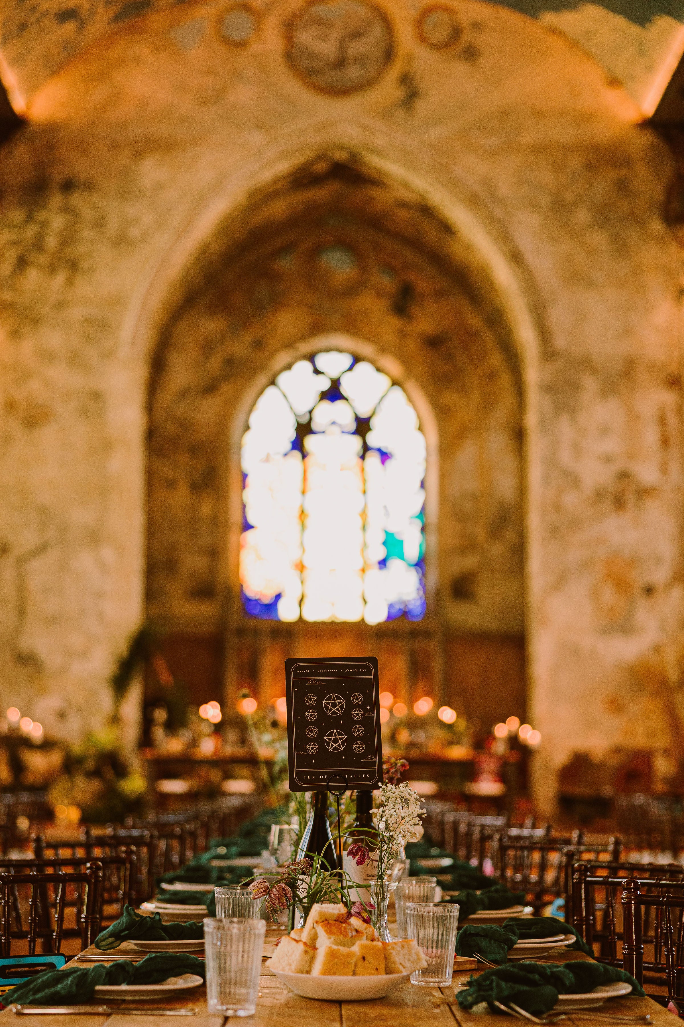 Luxury wedding stationery adorns a long table in a dramatic, vintage hall with gothic arches and a vivid stained glass window.