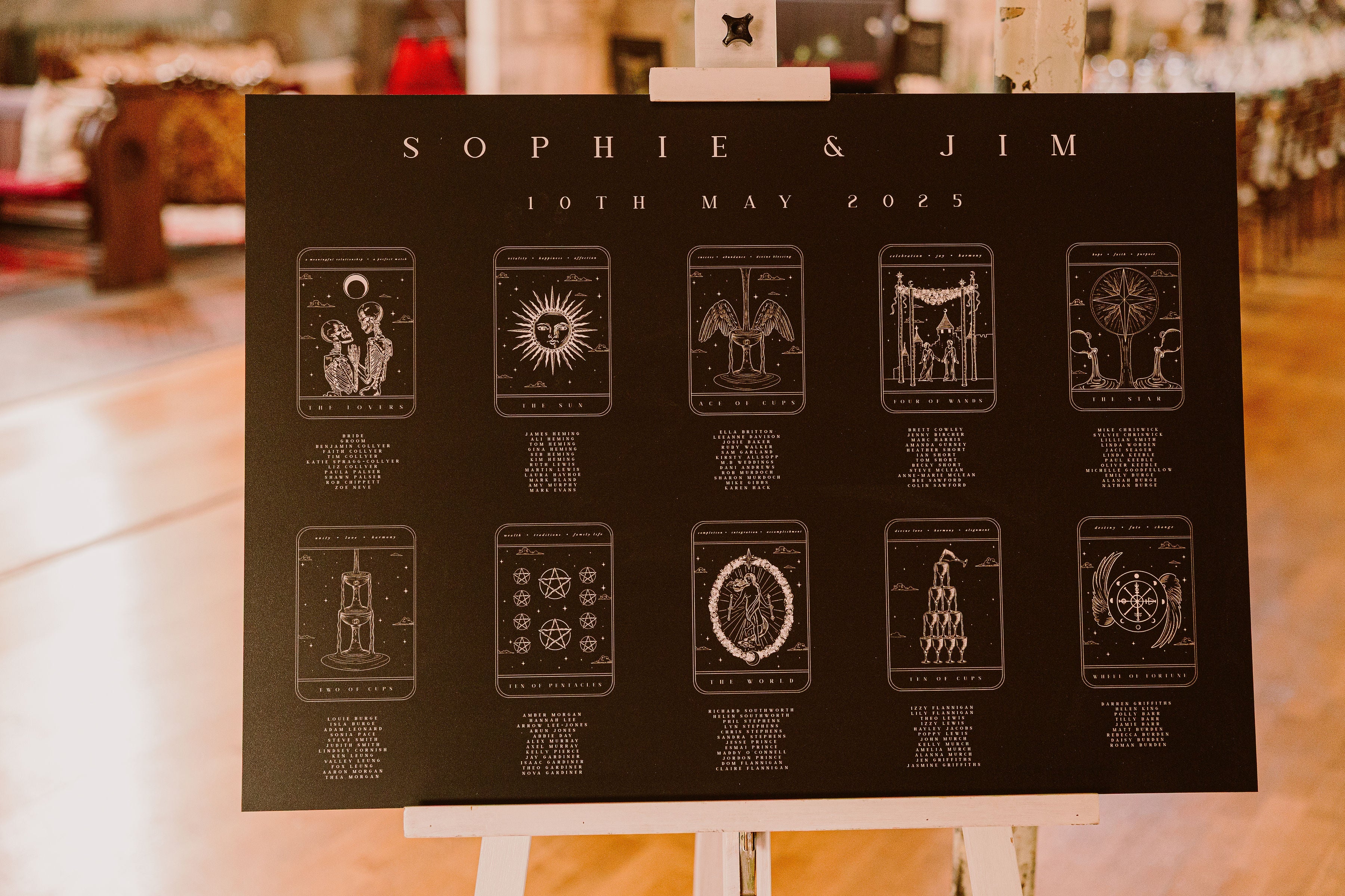 Luxury black wedding seating chart with mystical tarot-inspired design on an easel; dramatic, warmly lit UK venue backdrop.