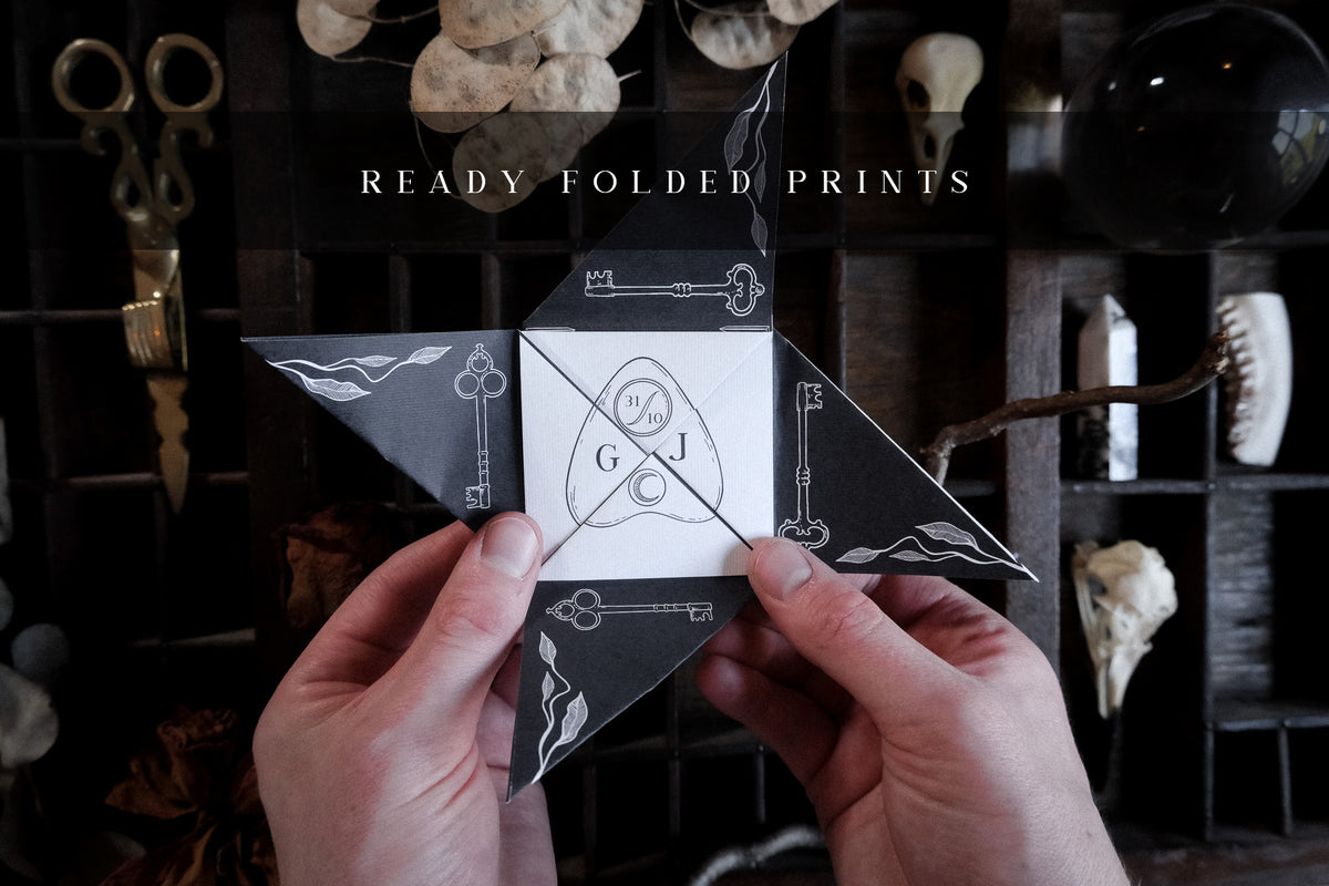 A black and white star-shaped Victorian folding wedding invitation features hand illustrations, keys and wave motifs, shown amidst vintage objects.